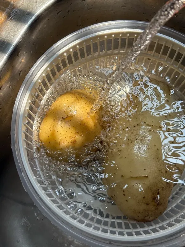 Two potatoes being rinsed under running water in a kitchen sink — illustrating how pasta or potato water can be reused as a mild fertilizer for Calathea plants.